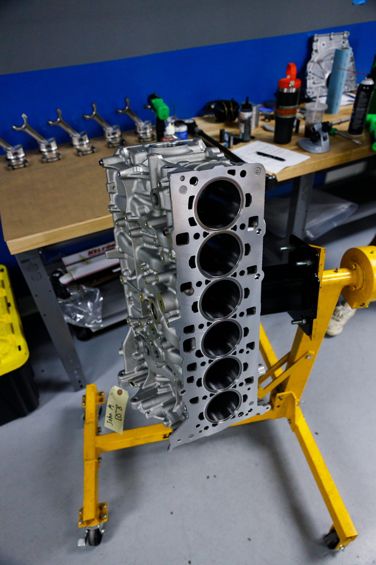 A silver aluminum engine block with four visible cylinder bores, mounted on a yellow engine stand in a workshop setting with a blue wall and workbench in the background.