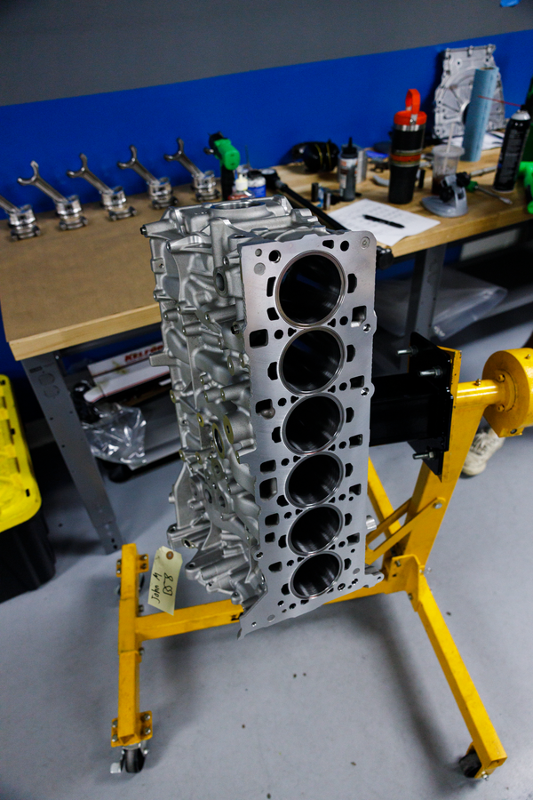 A silver aluminum engine block with four visible cylinder bores, mounted on a yellow engine stand in a workshop setting with a blue wall and workbench in the background.