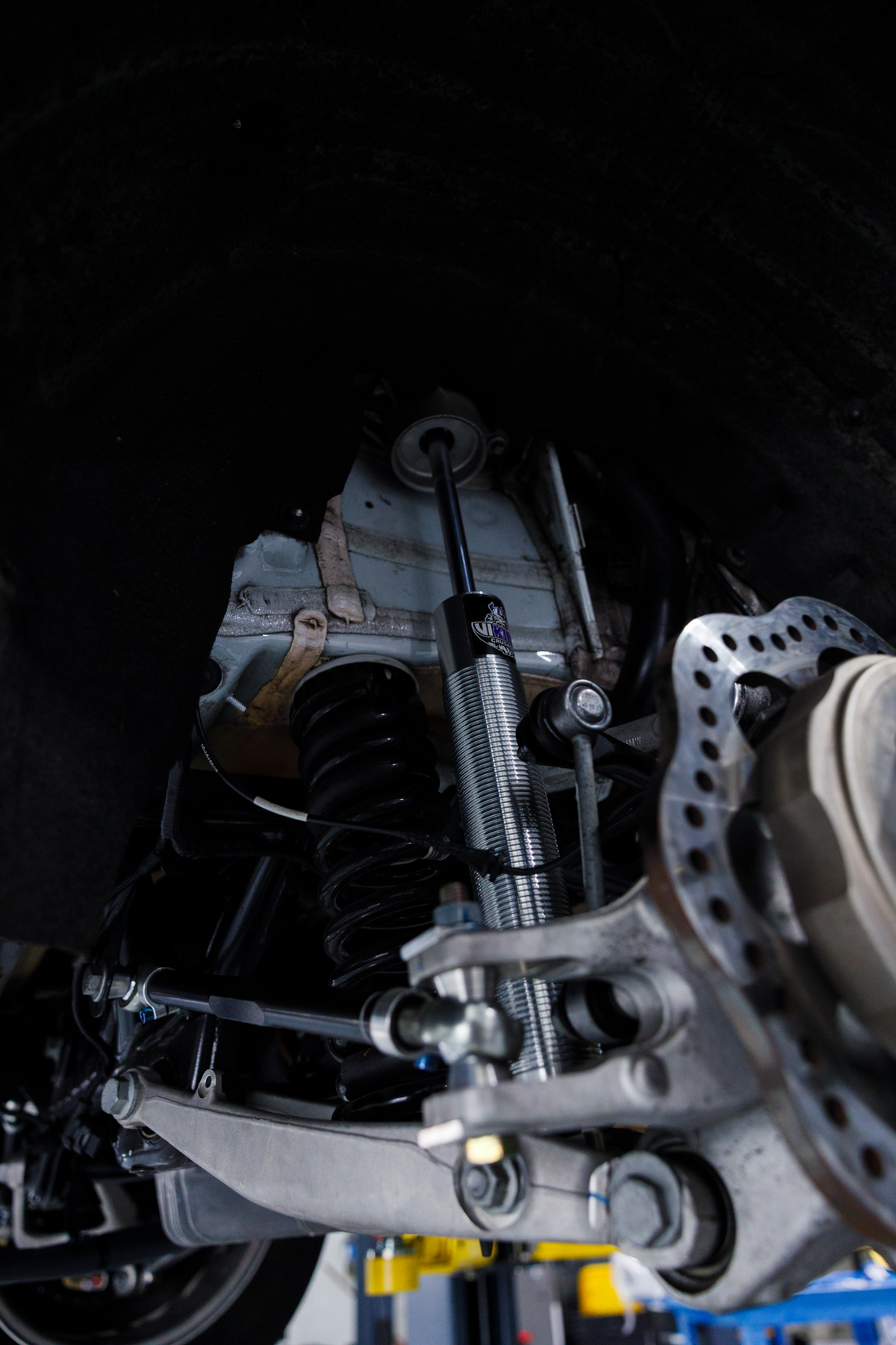 Close-up of a vehicle's undercarriage showing the suspension system with a shock absorber, coil spring, brake rotor, and control arms.